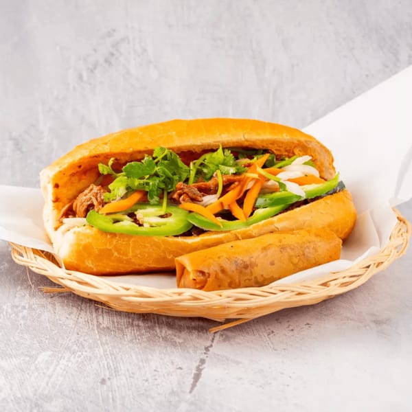 A Vietnamese banh mi sandwich with an egg roll in a woven basket on a gray surface.