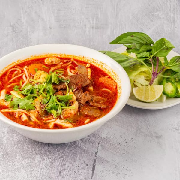 A bowl of ChuMinh Tofu's Vietnamese spicy beef noodle soup with a side of fresh herbs.